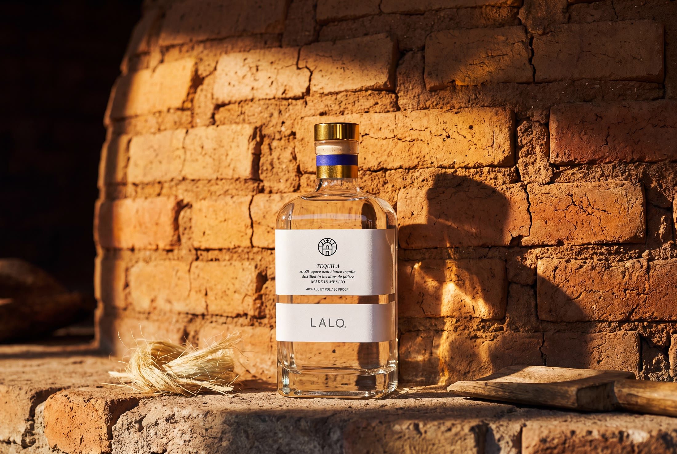 LALO blanco tequila bottle with a gold cap standing on a stone ledge against a sunlit adobe brick oven, agave fibres at its base — AI product photography by Chronos Studio.