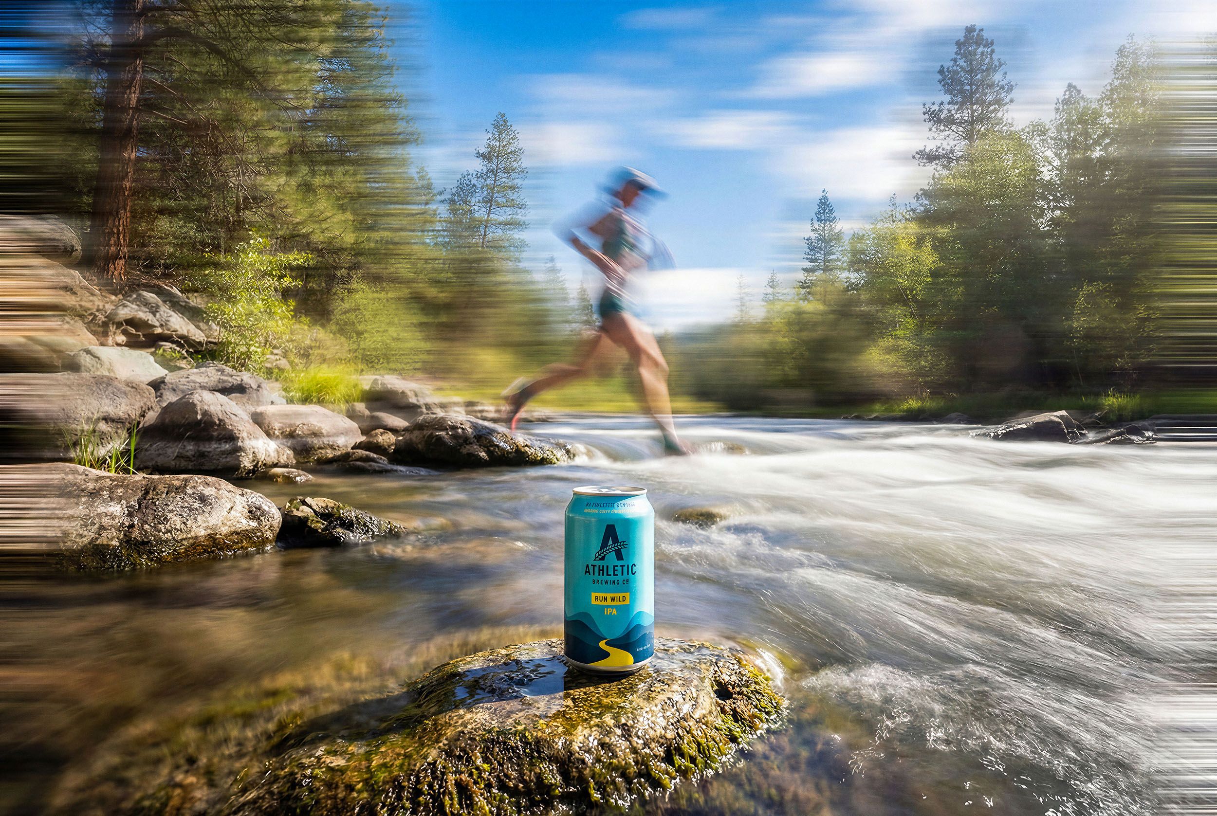 An Athletic Brewing 'Run Wild IPA' can on a river rock as a motion-blurred trail runner leaps across a sunlit forest stream.