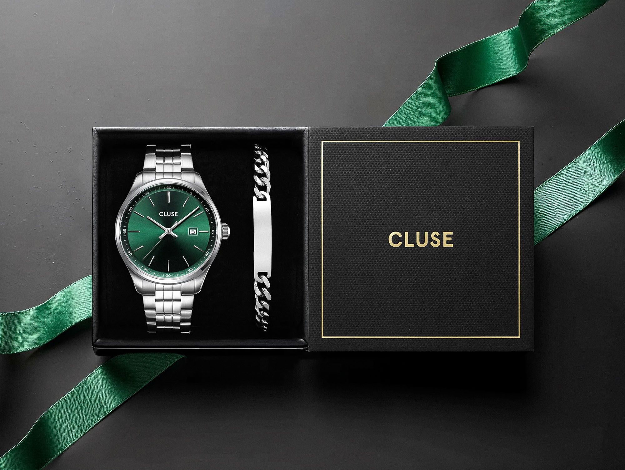 A CLUSE watch and steel bracelet presented in a black gift box with gold-foil logo, wrapped in green ribbon — premium product packaging.