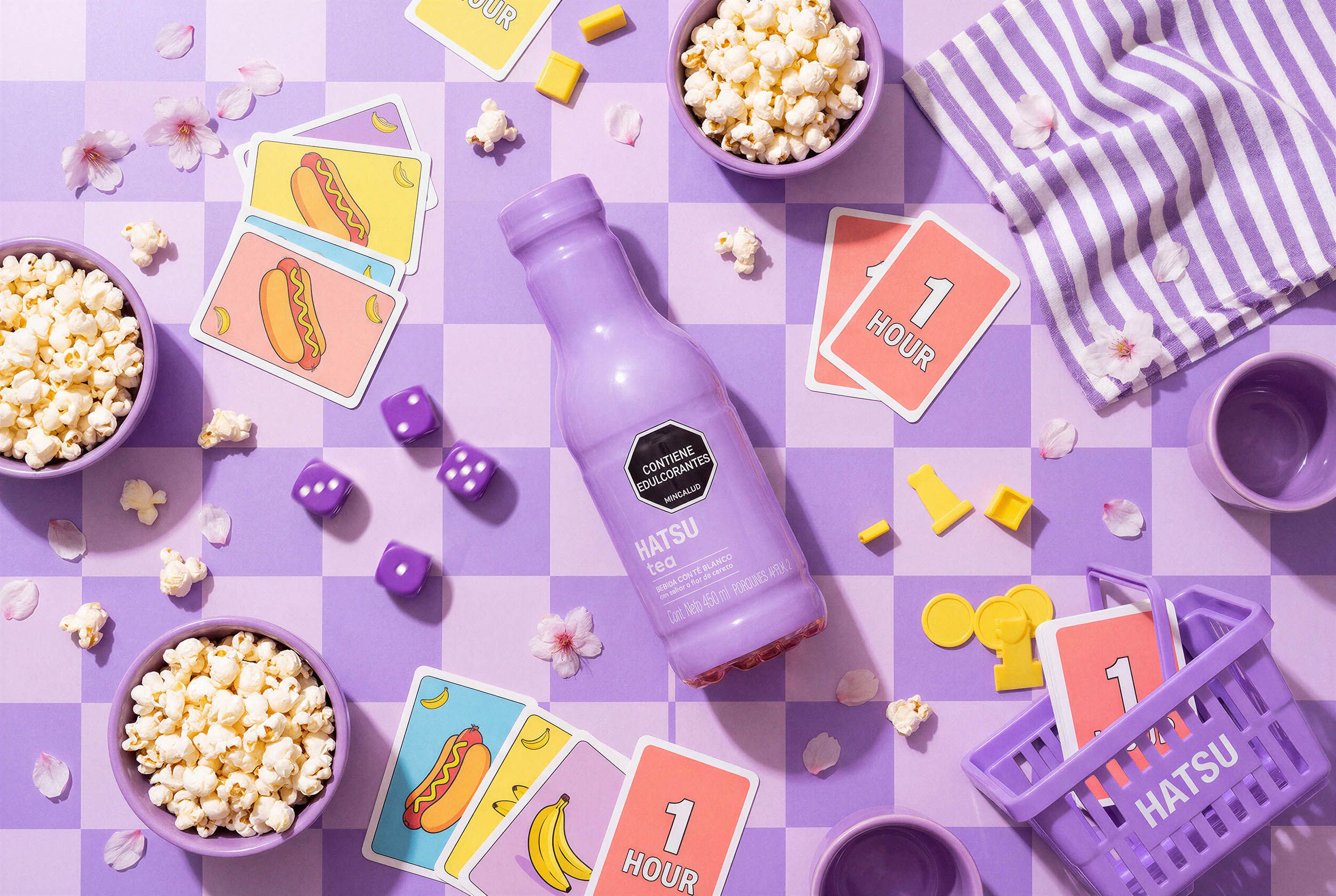 A playful lilac flat-lay of a Hatsu tea bottle surrounded by popcorn bowls, illustrated playing cards, dice and cherry blossoms on a purple checkerboard.