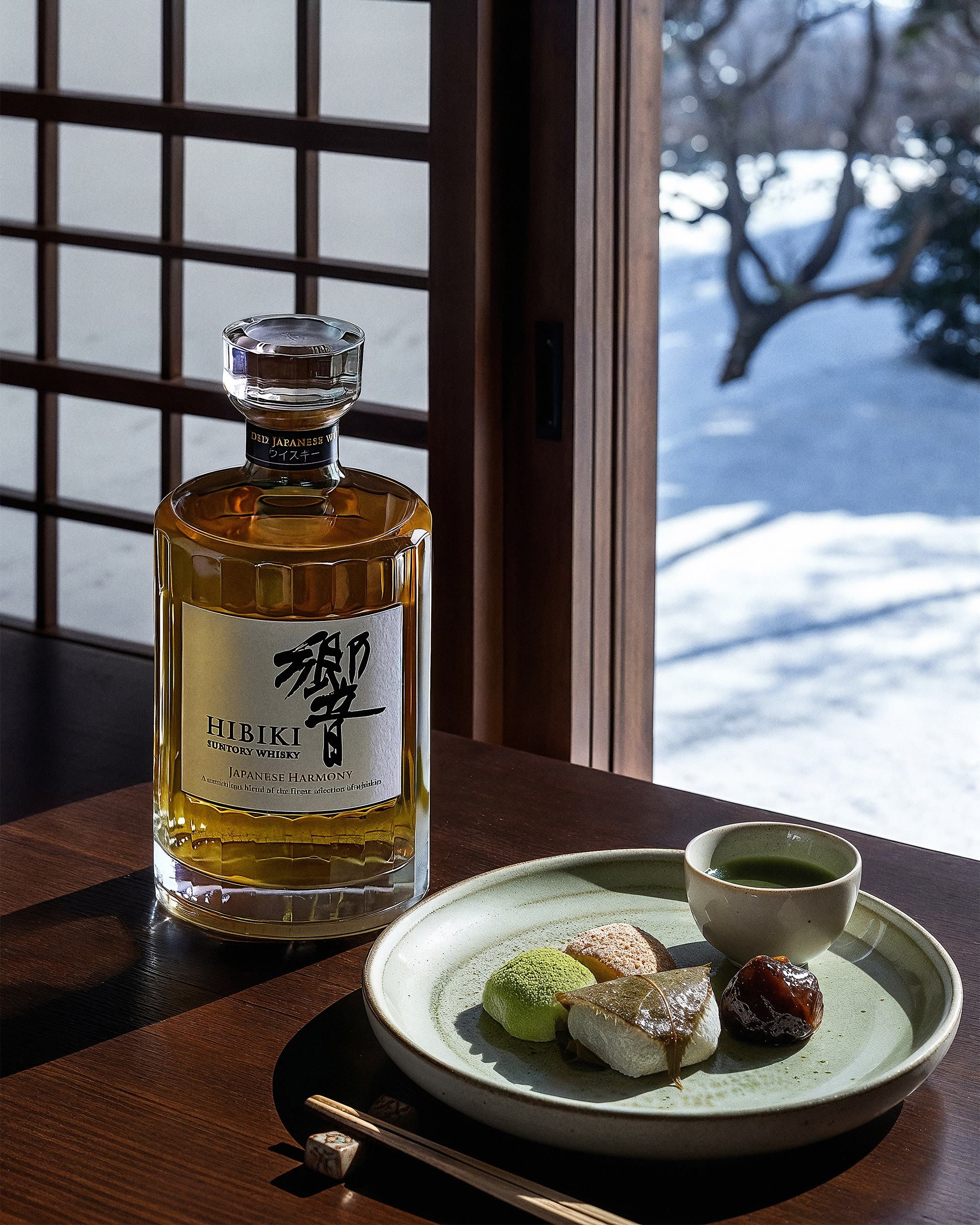 Hibiki bottle beside a plate of wagashi and matcha, a snowy Japanese garden beyond the window — Chronos Studio.