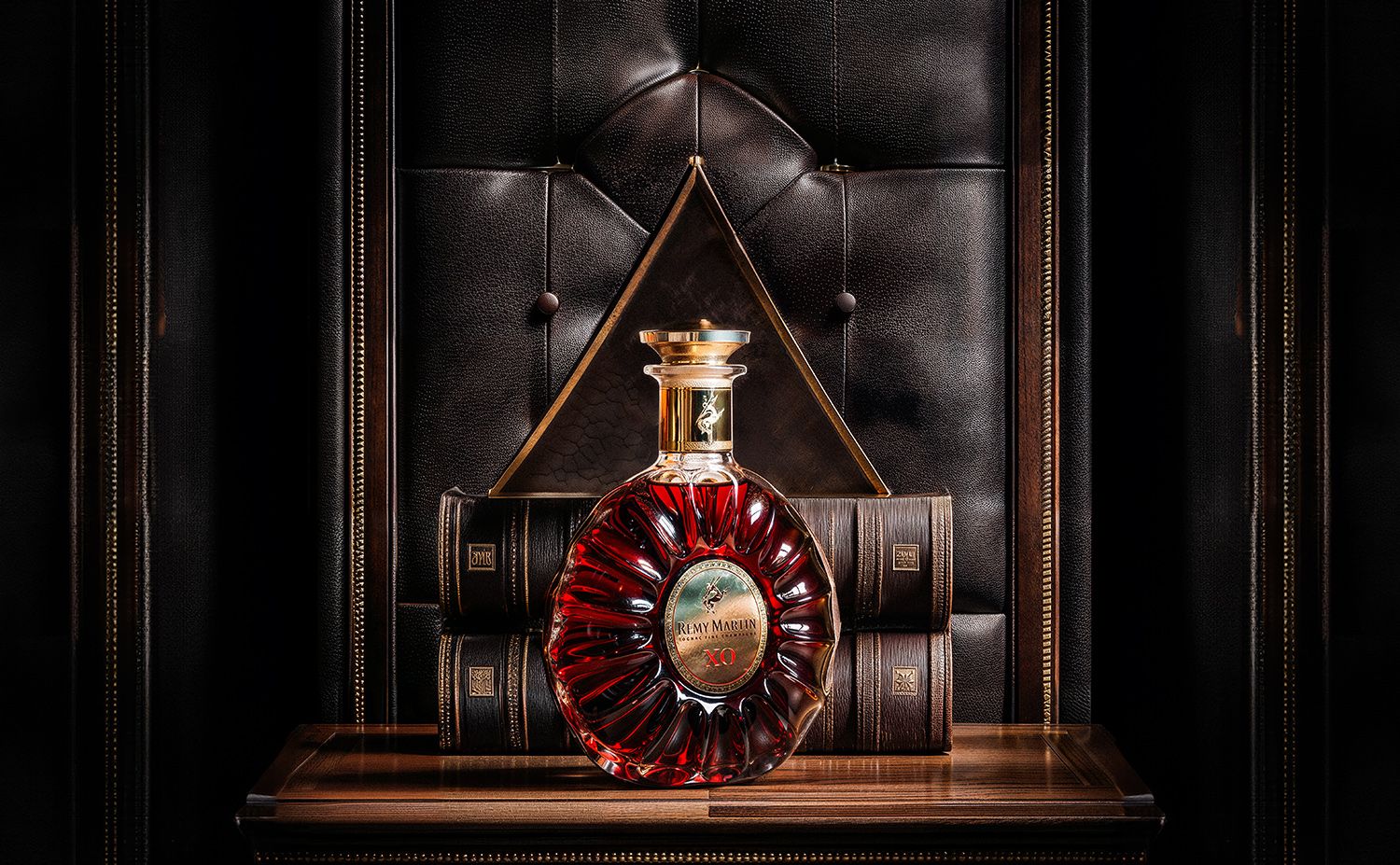 Rémy Martin XO decanter glowing ruby-red against tufted leather and antique books — dramatic still life by Chronos Studio.