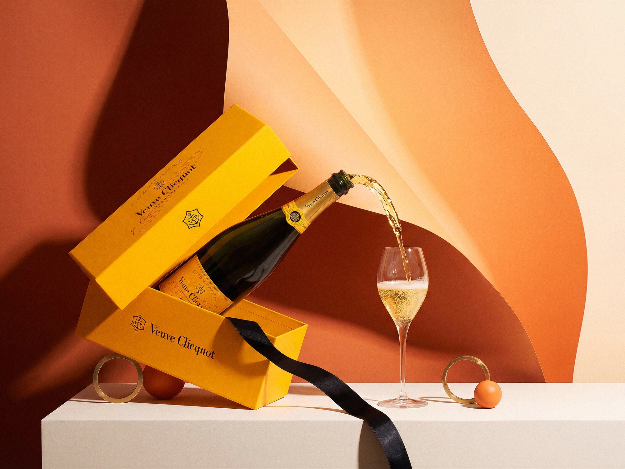 Veuve Clicquot Champagne pouring mid-air from a tilted bottle into a flute, yellow gift boxes stacked on sculpted terracotta paper waves — holiday product photography by Chronos Studio.