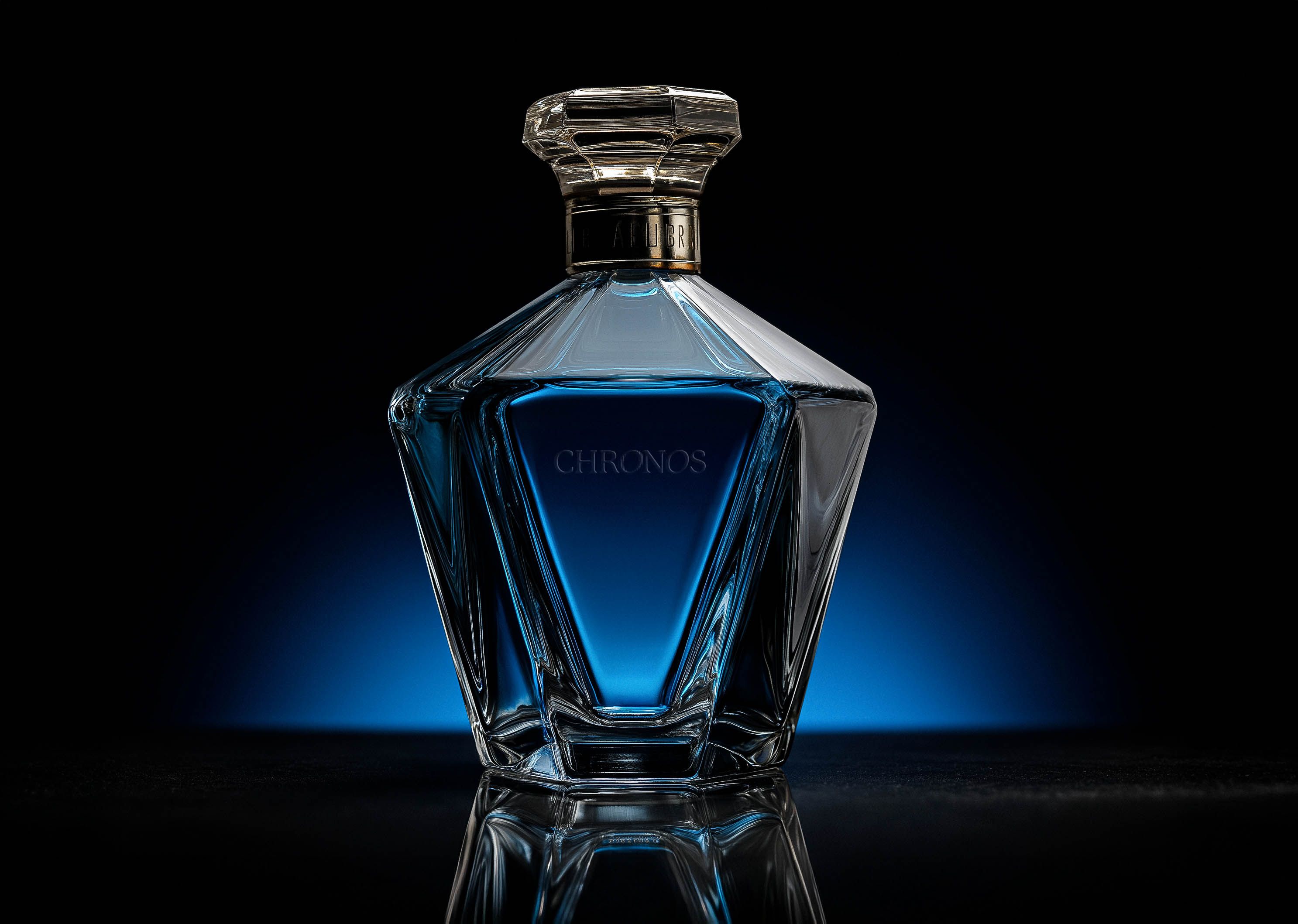 A faceted crystal decanter of blue spirit engraved 'CHRONOS', lit with a dramatic blue glow on a black reflective surface.