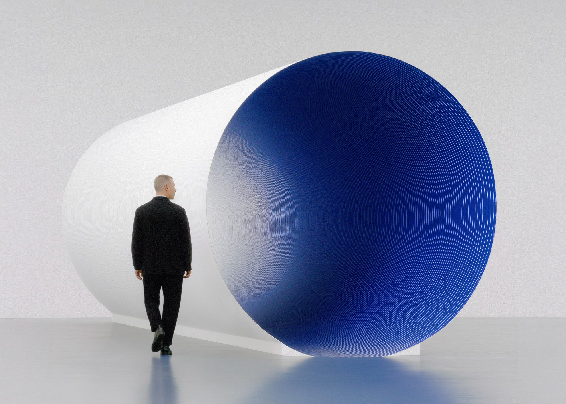 A man in a black suit walking past a monumental white cylinder with a deep cobalt-blue circular face in a bright minimalist gallery.