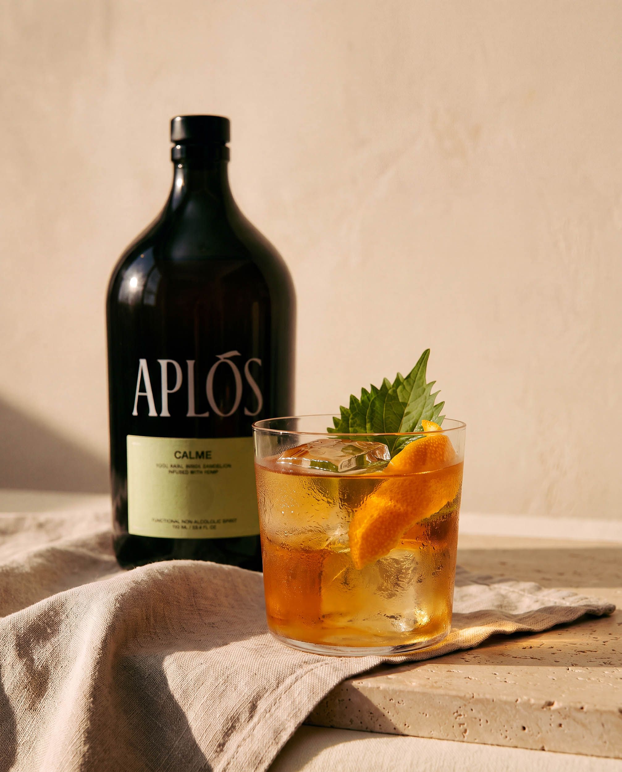 The Serve — macro of a chilled lowball with hand-cut ice, mandarin peel and a shiso leaf, condensation catching low golden sun, the Aplós bottle soft behind.