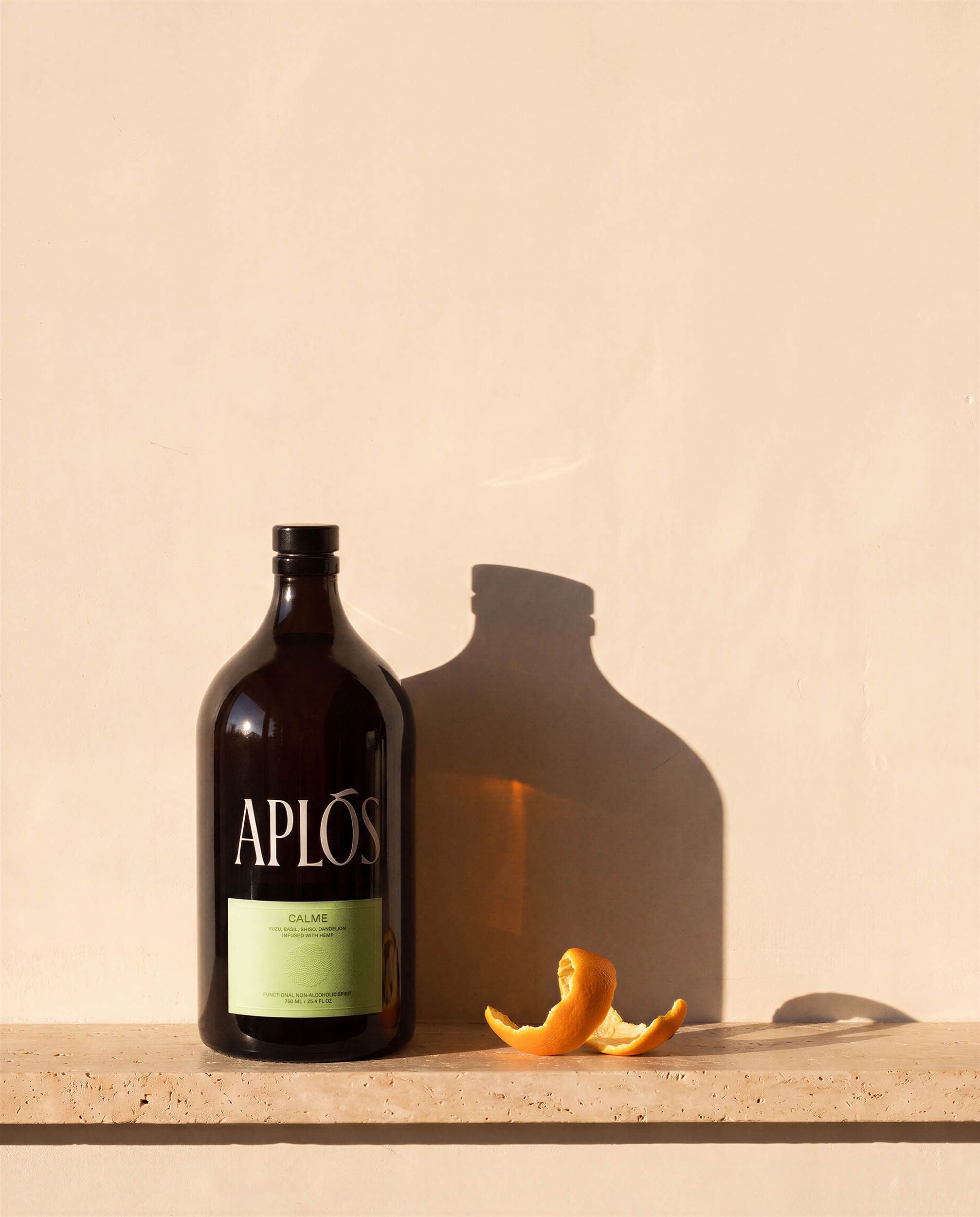 The Stillness — the Aplós bottle alone on a travertine ledge, casting one long clean shadow across a bone-cream plaster wall with vast calm negative space above.